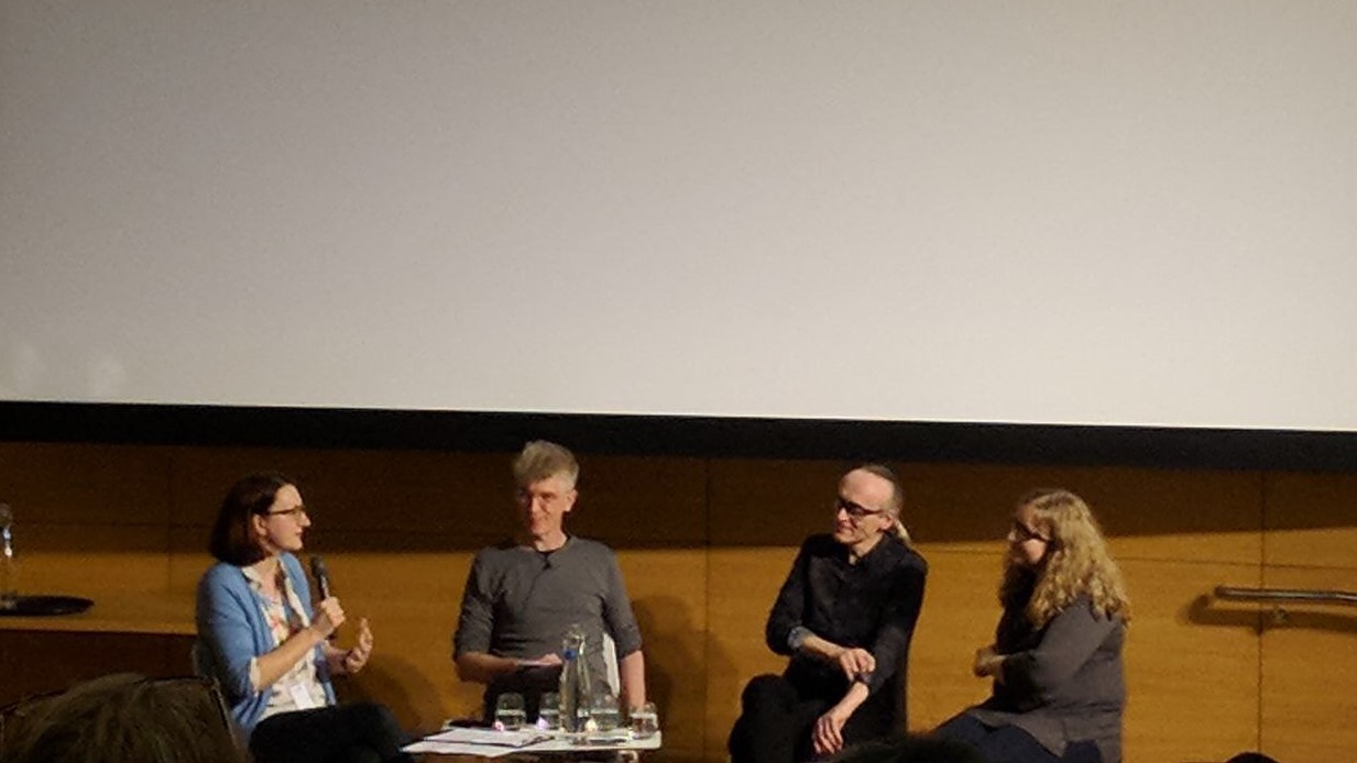 Four people sitting on a stage infront of a big screen.